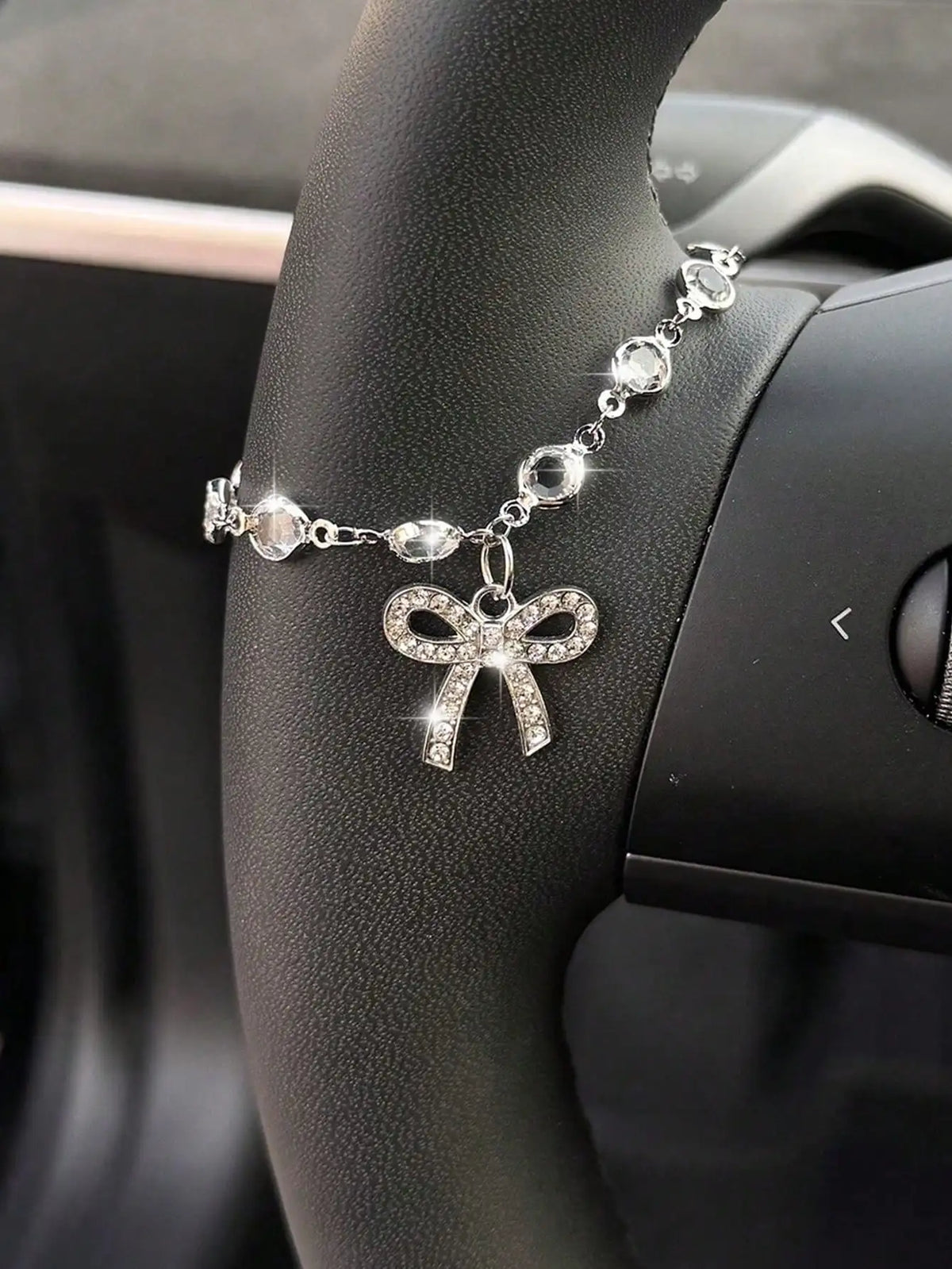 A silver rhinestone bow cross pendant necklace worn on a person's wrist, set against a dark car interior.