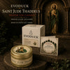 Evoduck Saint Jude Thaddeus prayer tin candle and guide set displayed on a dark surface with a religious statue in the backgr