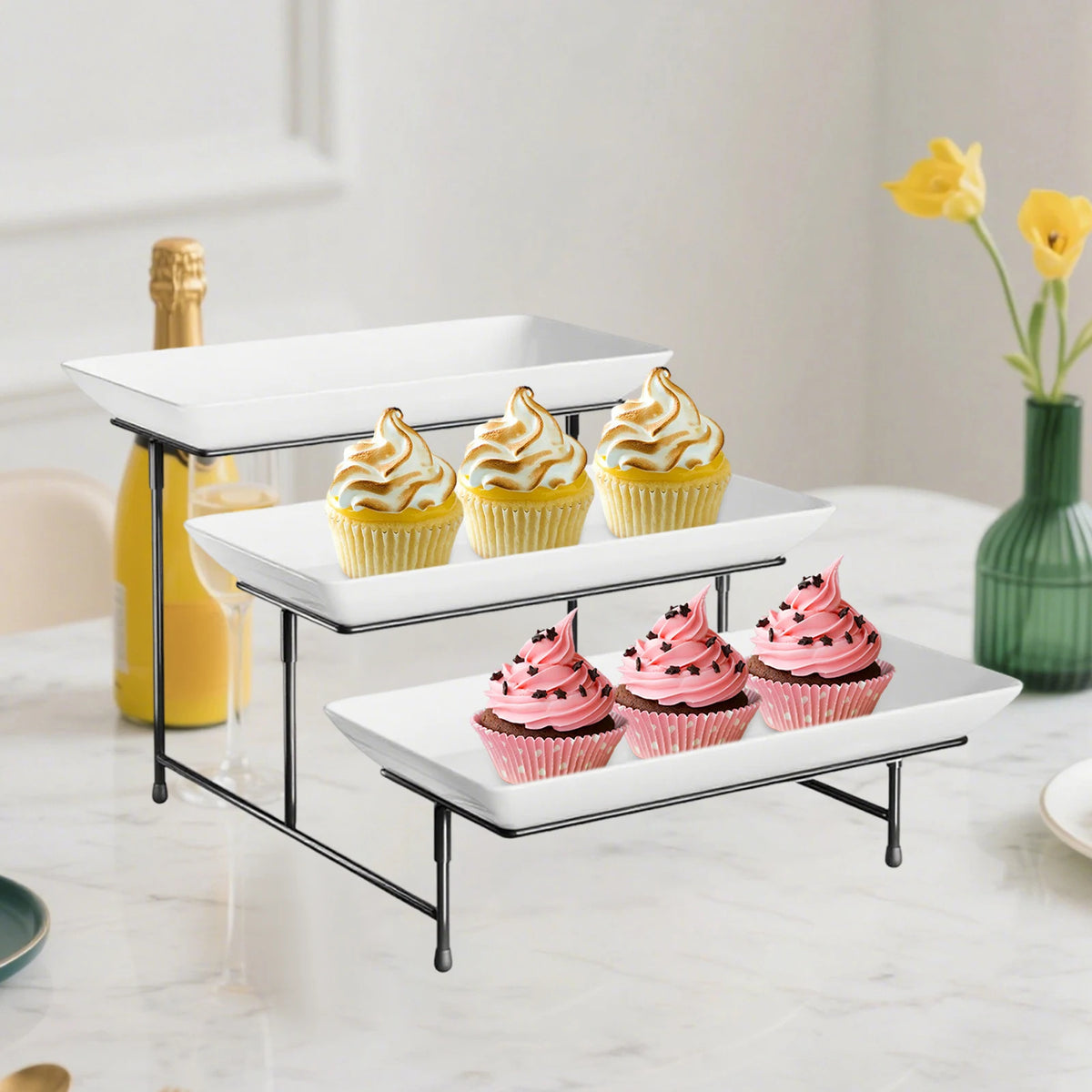 A black metal 3-tier rectangular serving platter stand holding cupcakes and a bottle of yellow liquid.