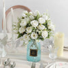 A bouquet of white artificial roses in a light blue glass vase with greenery on a white table.