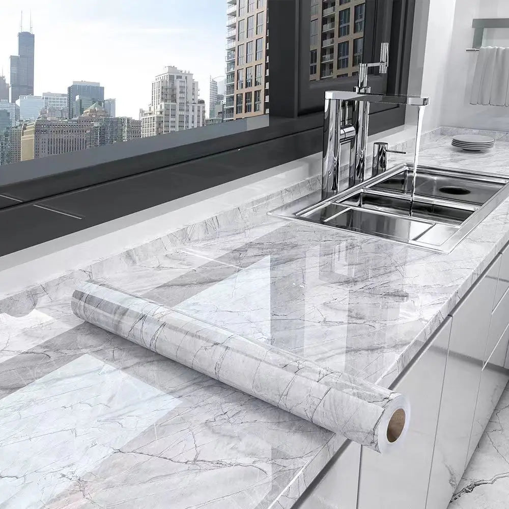 A stainless steel kitchen sink and faucet are installed in a white marble countertop, with a roll of marble self-adhesive wal