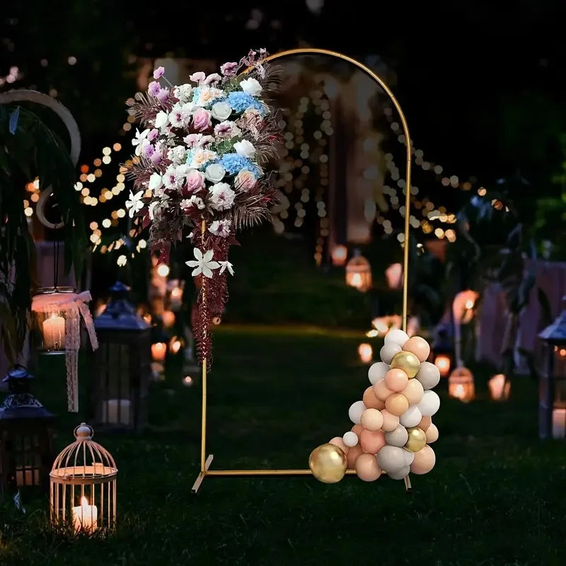 A gold metal arch stand decorated with floral arrangements and string lights set up outdoors for an event.