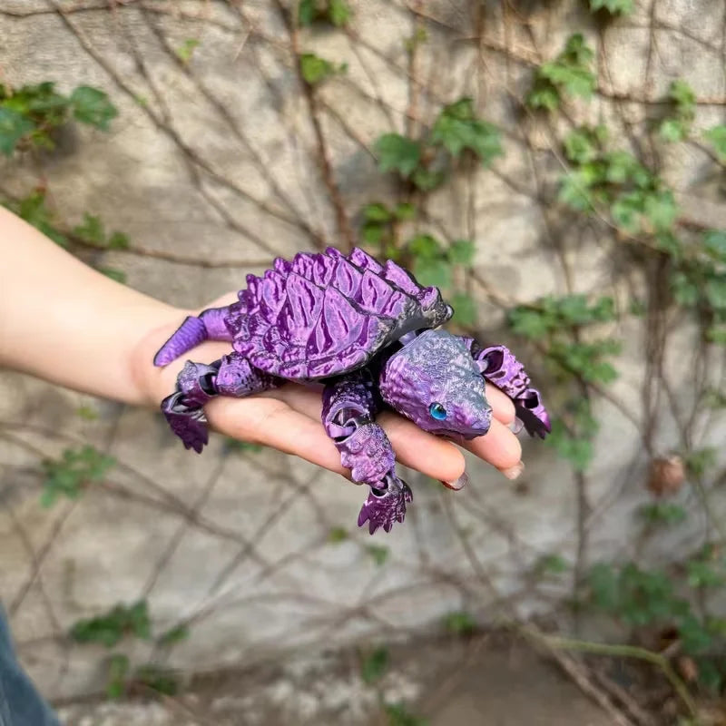 A purple 3D printed alligator snapping turtle figurine with articulated joints is held in a person's hand against a blurred g