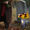A person in a brown coat holds flowers near a stone grave marker with an angel statue and a cross memorial vase.