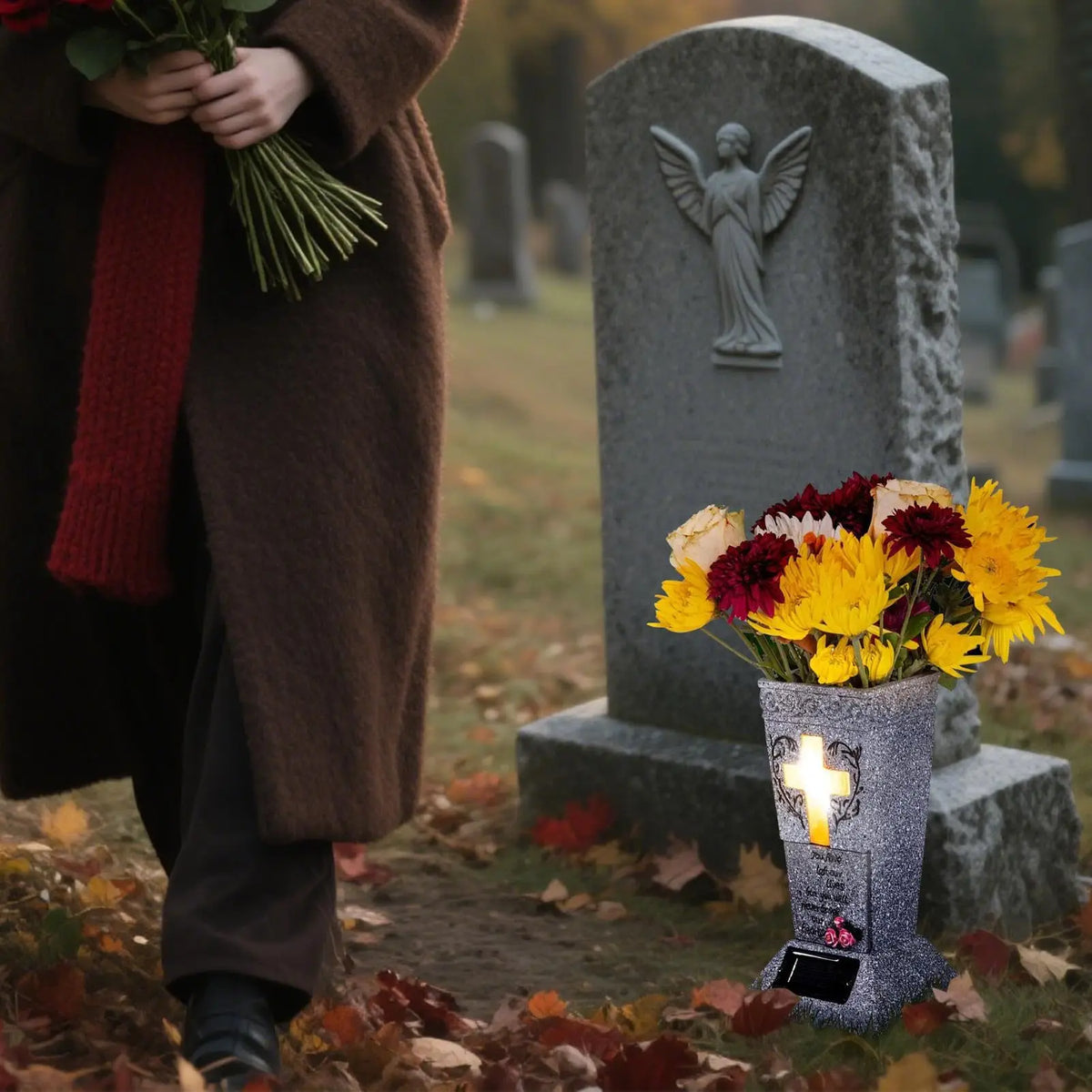 A person in a brown coat holds flowers near a stone grave marker with an angel statue and a cross memorial vase.