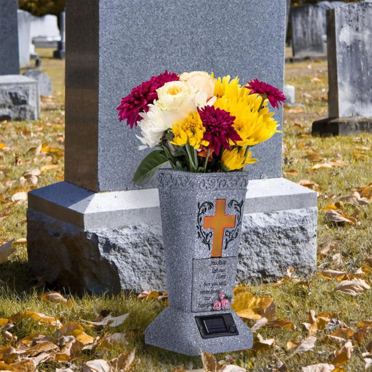 A gray solar LED cross memorial vase placed on a stone grave marker in a cemetery.