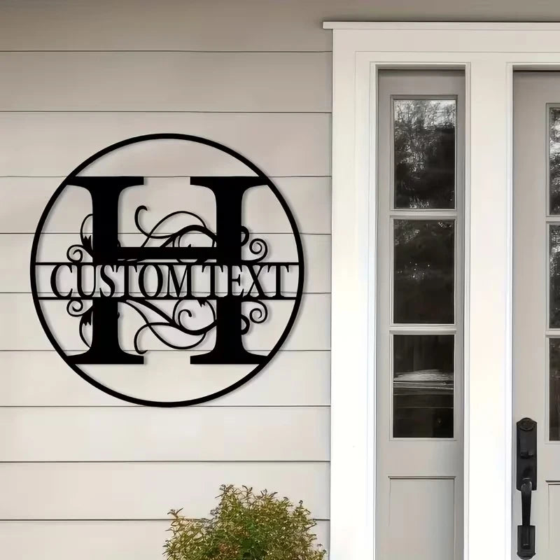A black metal initial sign with custom text is mounted on a white clapboard siding next to a white door frame.