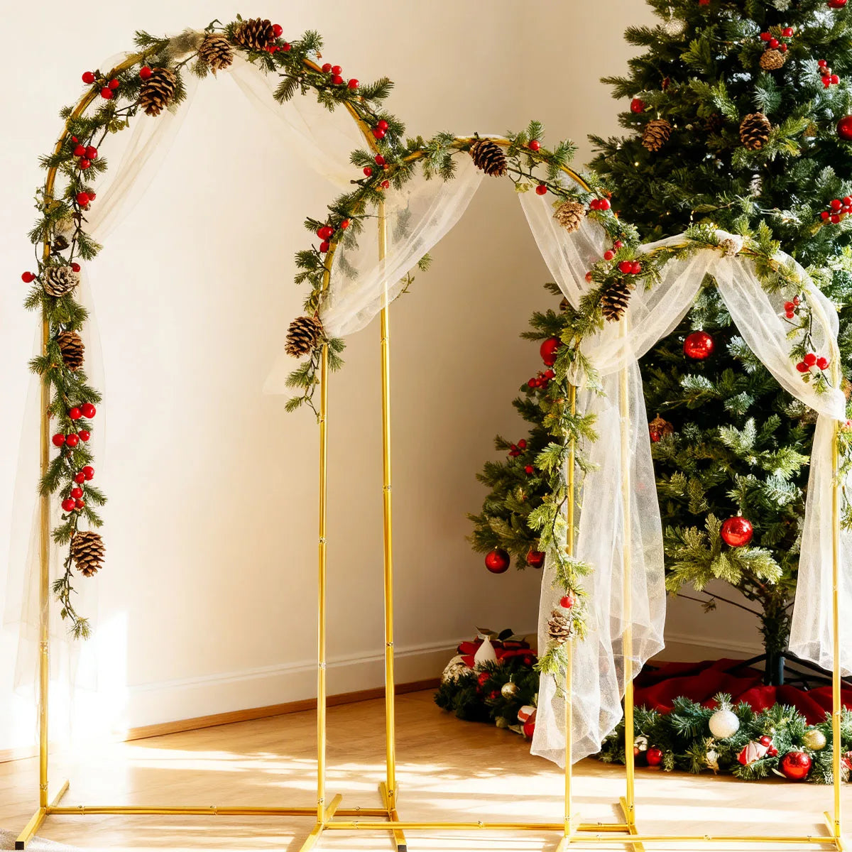 A gold metal arch backdrop decorated with greenery, pinecones, and red berries, draped with sheer white fabric.
