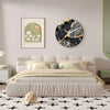 A modern marble textured glass wall clock with a white face and gold accents hangs above a beige upholstered bed.
