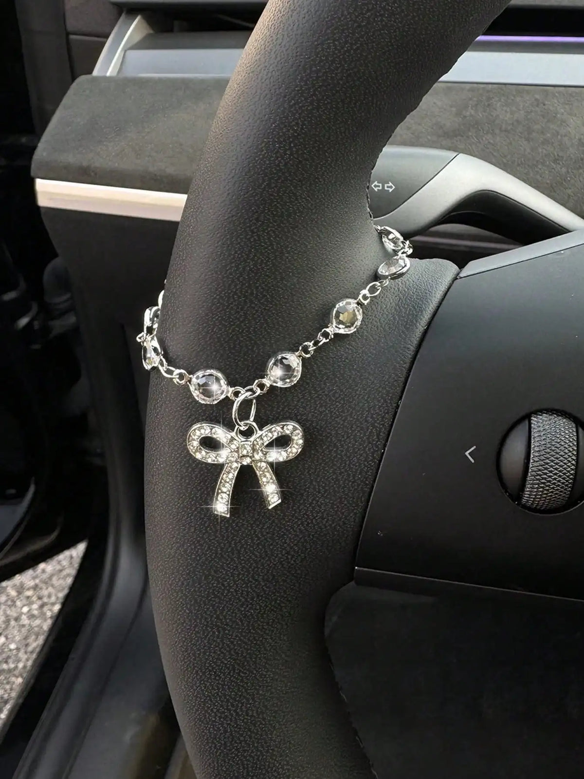 A silver rhinestone bow cross pendant necklace is displayed on a person's hand next to a car steering wheel.