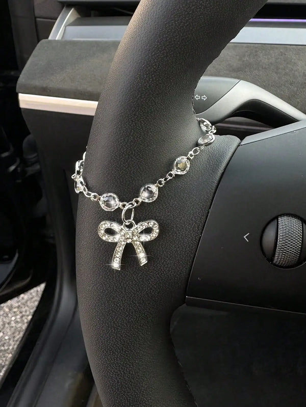 Un pendentif en forme de nœud de strass argenté est exposé sur la main d'une personne à côté d'un volant de voiture.