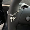 A silver rhinestone bow cross pendant necklace is displayed on a person's hand next to a car steering wheel.