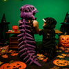 Peluche squelette de dinosaure violet devant une figure de sorcière noire et des citrouilles sculptées.