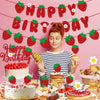 A festive birthday banner with strawberries and the words HAPPY BIRTHDAY, surrounded by various strawberry-themed party suppl