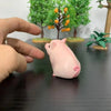 A pink mini pig figurine is being held by a hand, displayed in front of a green tree arrangement.