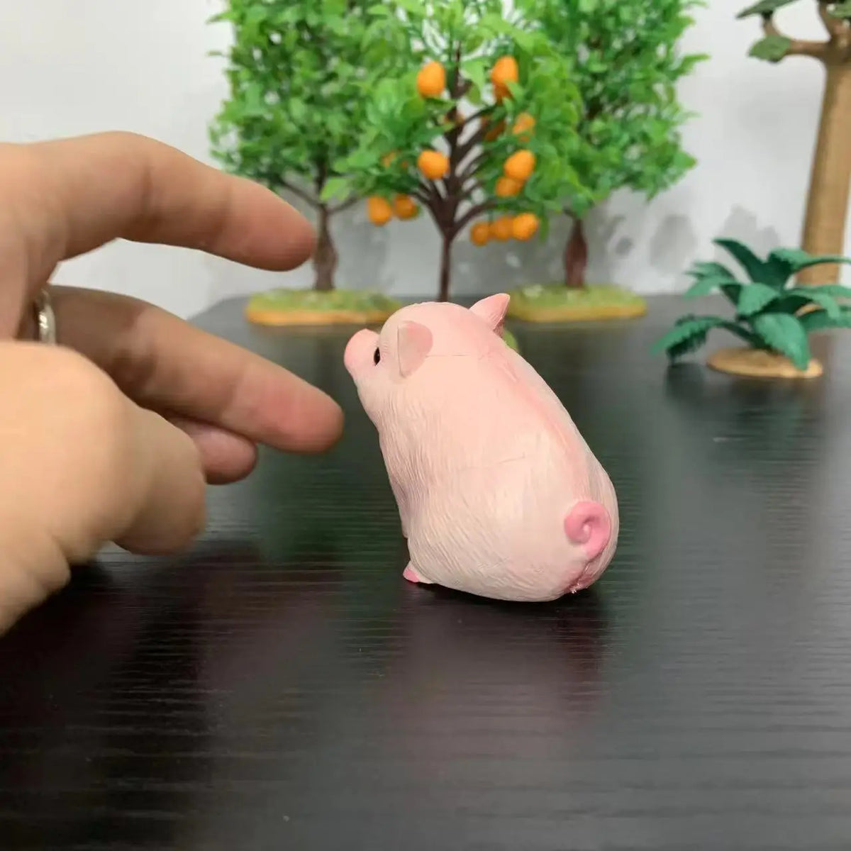 A pink mini pig figurine is being held by a hand, displayed in front of a green tree arrangement.
