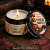 A black tin scented candle labeled Pumpkin Chai Doorstep resting on a dark wooden surface with autumn leaves.