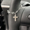 A silver rhinestone cross pendant necklace worn on a person's wrist next to a car steering wheel.