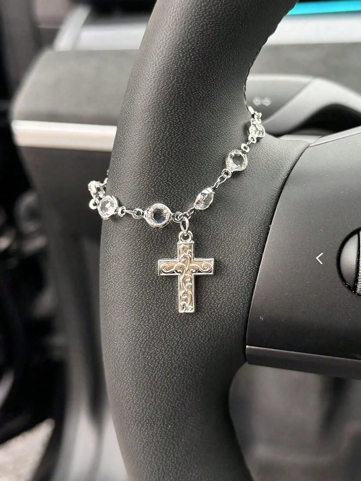 A silver rhinestone cross pendant necklace worn on a person's wrist next to a car steering wheel.