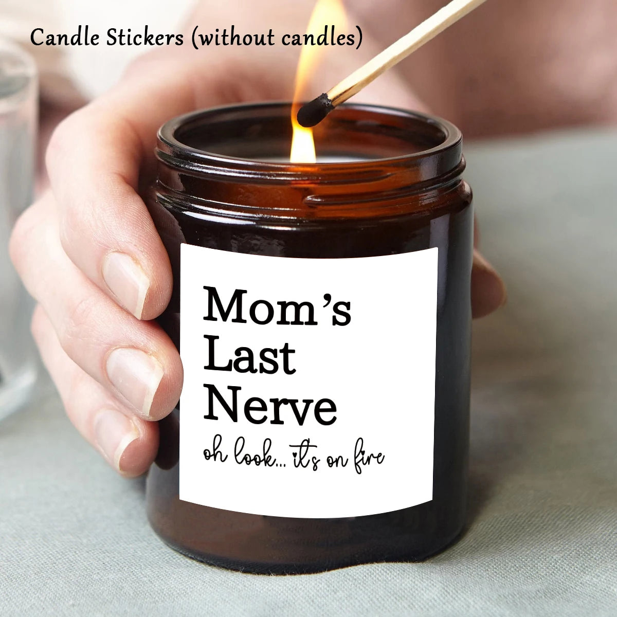 A hand lights a jar candle with a wick, showing a label that reads Mom's Last Nerve oh look it's on fire