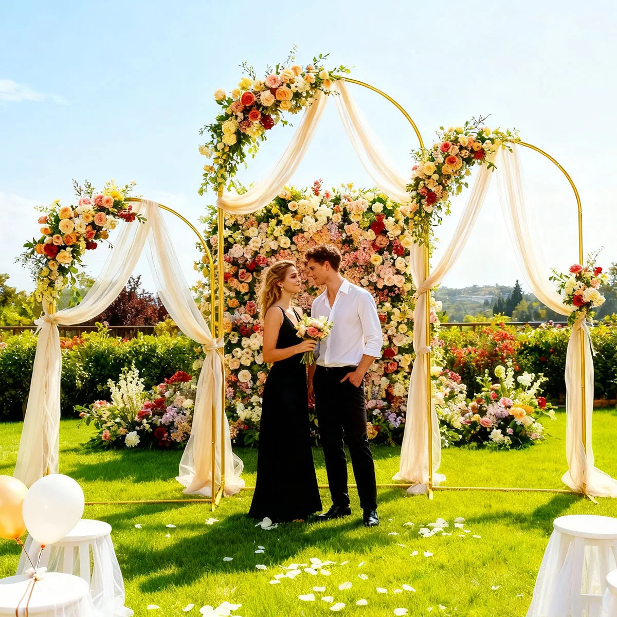 A bride and groom stand under a floral metal arch backdrop for a wedding photo shoot in a garden.
