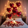 A bed decorated with red rose petals and heart-shaped foil balloons in a romantic setting.