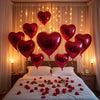 A bed decorated with red rose petals and heart-shaped foil balloons in various shades of pink and red.