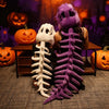Un peluche squelette de dinosaure violet à côté d'un modèle squelette blanc dans un décor d'Halloween.
