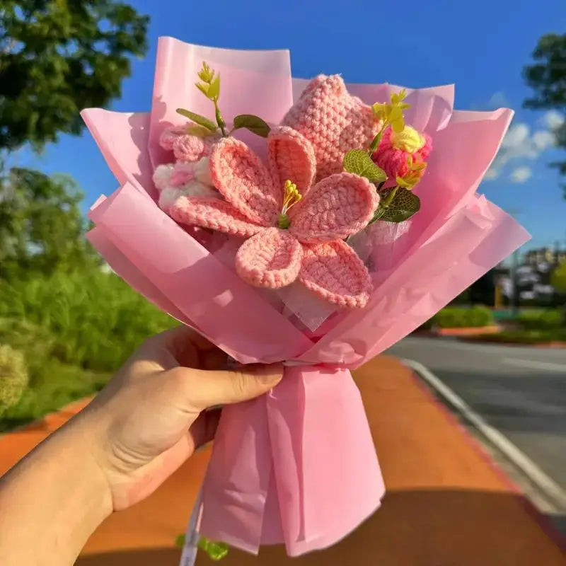 A pink handmade crochet yarn flower bouquet held by a hand outdoors against a blue sky background.