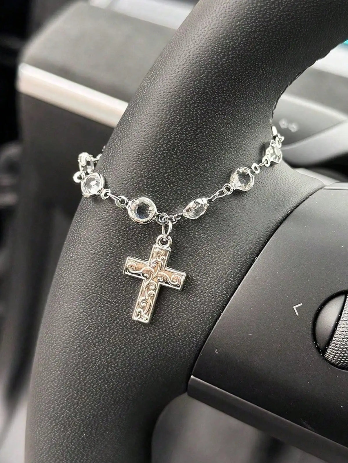 Silver rhinestone cross pendant necklace worn on a wrist, set against a dark car steering wheel.
