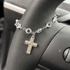 Silver rhinestone cross pendant necklace worn on a wrist, set against a dark car steering wheel.