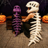 Une personne pose avec deux peluches squelettes de dinosaures réalistes, un violet et un blanc, sur fond sombre.
