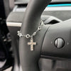 A silver rhinestone bow cross pendant necklace is displayed on a person's wrist next to a car steering wheel.