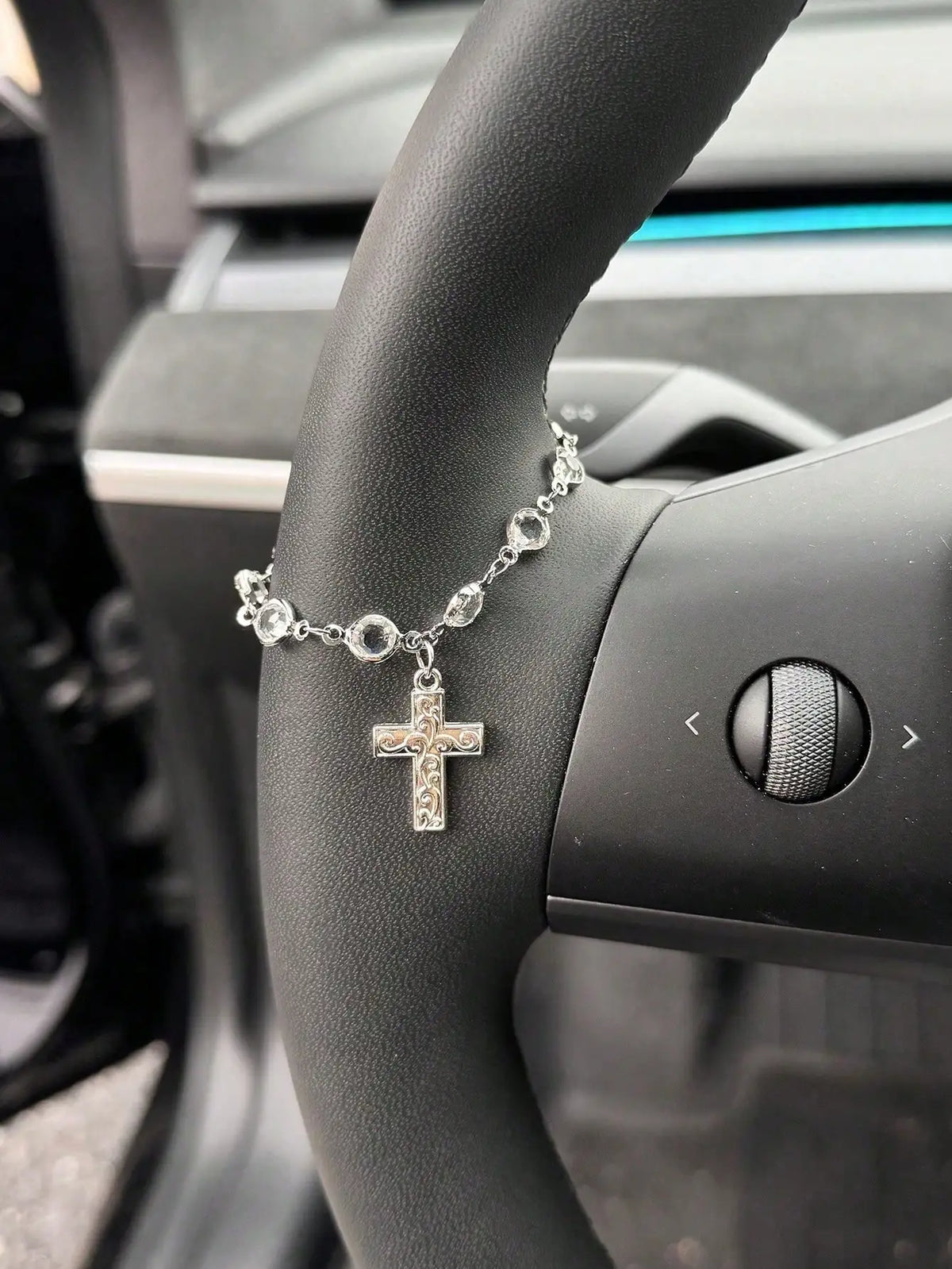 A silver rhinestone bow cross pendant necklace is displayed on a person's wrist next to a car steering wheel.