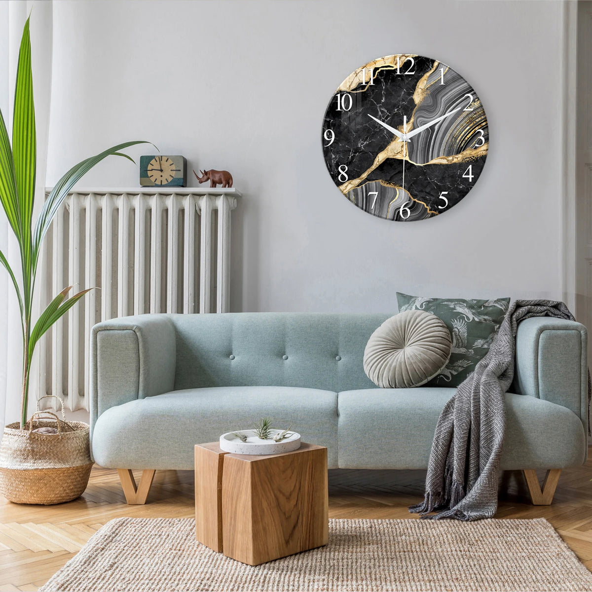 A modern marble textured glass wall clock with gold accents hangs on a light gray wall above a light blue sofa.