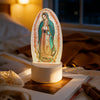 Acrylic LED night light of Our Lady of Guadalupe with a green and pink halo, resting on a wooden surface.