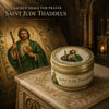 A prayer candle for Saint Jude Thaddeus in a decorative tin set on an ornate surface.