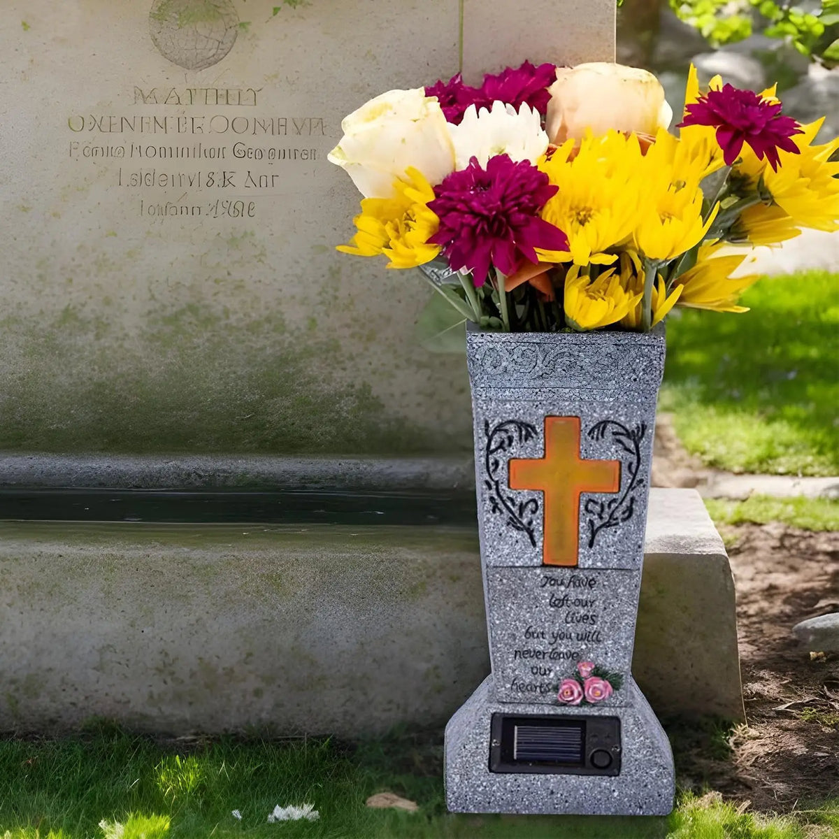 A gray memorial vase with a solar LED cross and yellow and purple flowers placed in front of a stone plaque.