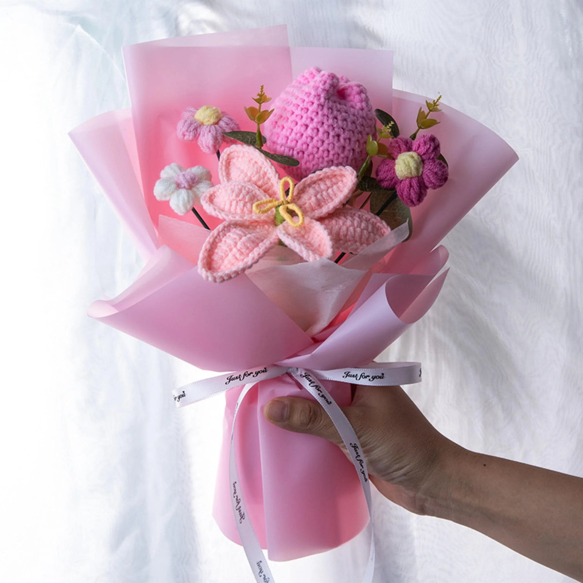 A bouquet of handmade crochet yarn flowers in pink and purple nestled in sheer pink paper.