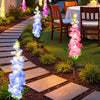 A garden pathway lined with pink and blue illuminated delphinium flower lights leading to a dining table.