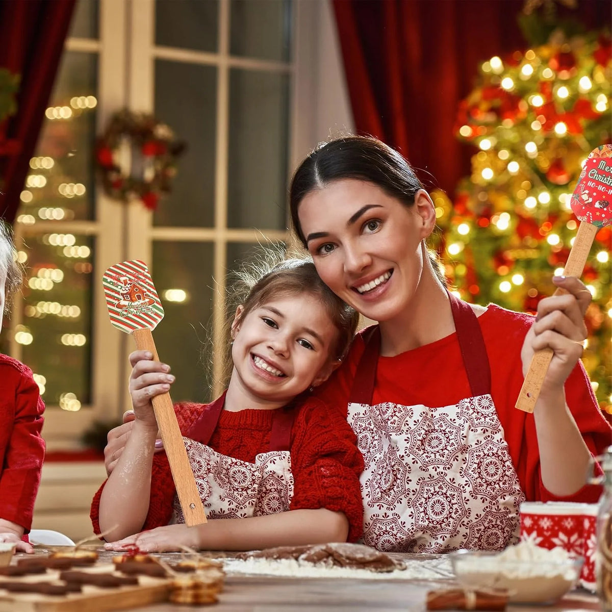A woman is holding a festive Christmas silicone spatula with a wooden handle while smiling and interacting with a young girl 