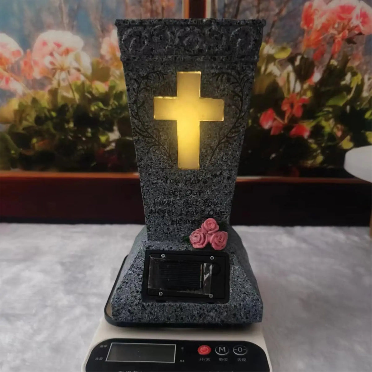 A dark gray memorial vase with a glowing yellow cross is displayed on a white electronic base.