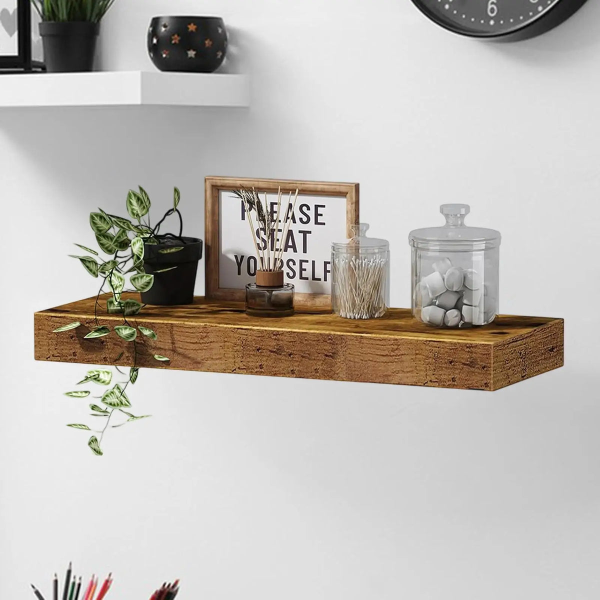 A rustic wooden floating shelf displaying a framed sign, candles, and glass jars on a white wall.