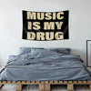 A black tapestry with the text Music Is My Drug in beige lettering hanging above a gray bed.
