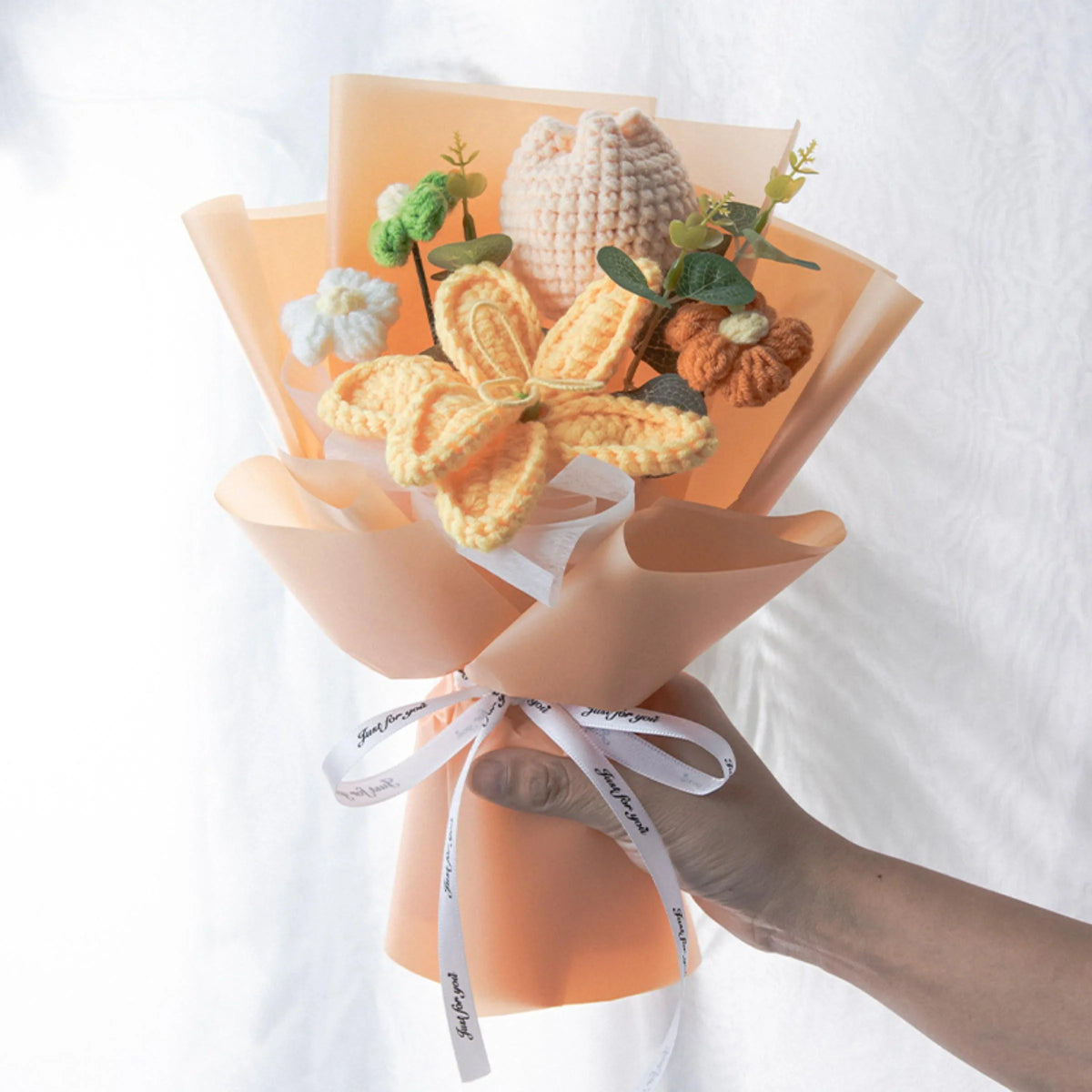A bouquet of handmade crochet yarn flowers in shades of yellow and orange arranged in a light peach paper wrapping.
