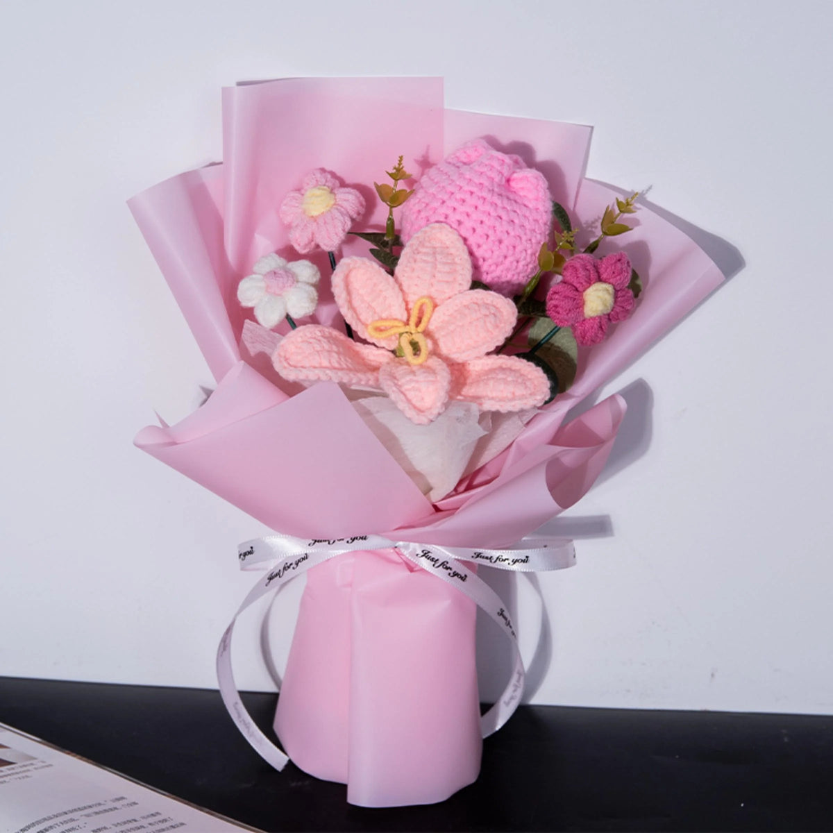 A bouquet of handmade crochet yarn flowers in pink and purple hues wrapped in sheer pink paper.