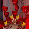 A display of red and gold heart-shaped balloons, candles, and red roses arranged for a special occasion.