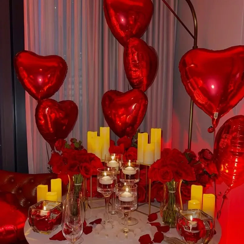 A display of red and gold heart-shaped balloons, candles, and red roses arranged for a special occasion.