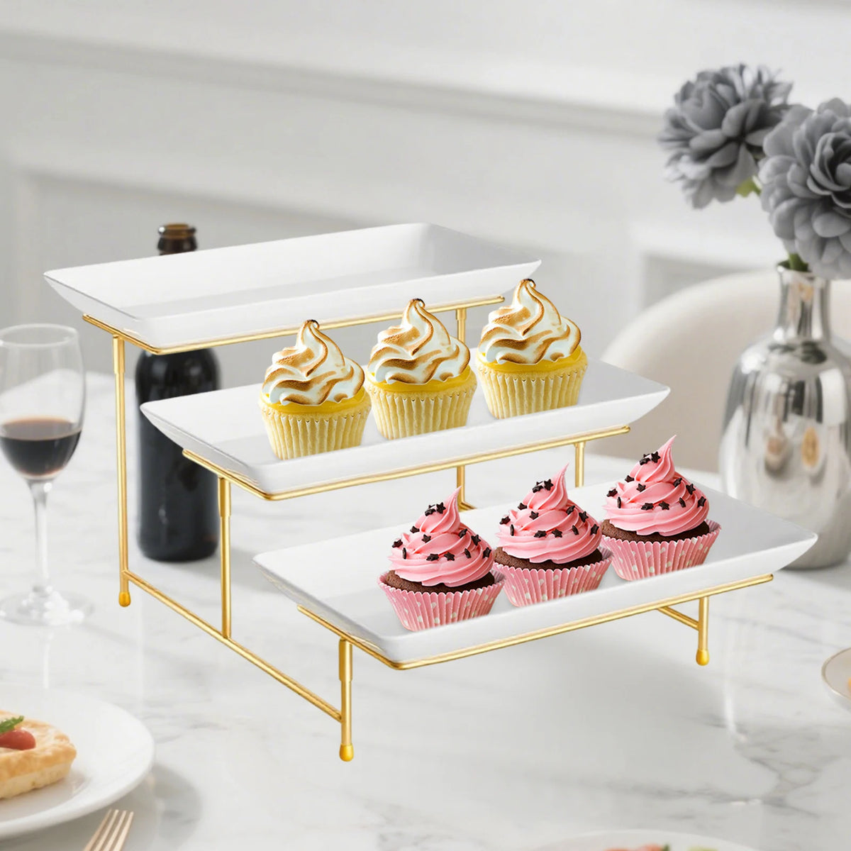 A gold-edged, three-tier rectangular serving platter stand displaying cupcakes and macarons on a white marble surface.