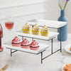 A three-tier rectangular serving platter stand holding cupcakes, featuring white and gold frosting on a modern black metal tr